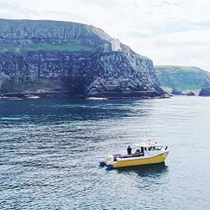 Small yellow-and-white motorboat with two people on calm blue water, with towering green cliffs in the background.